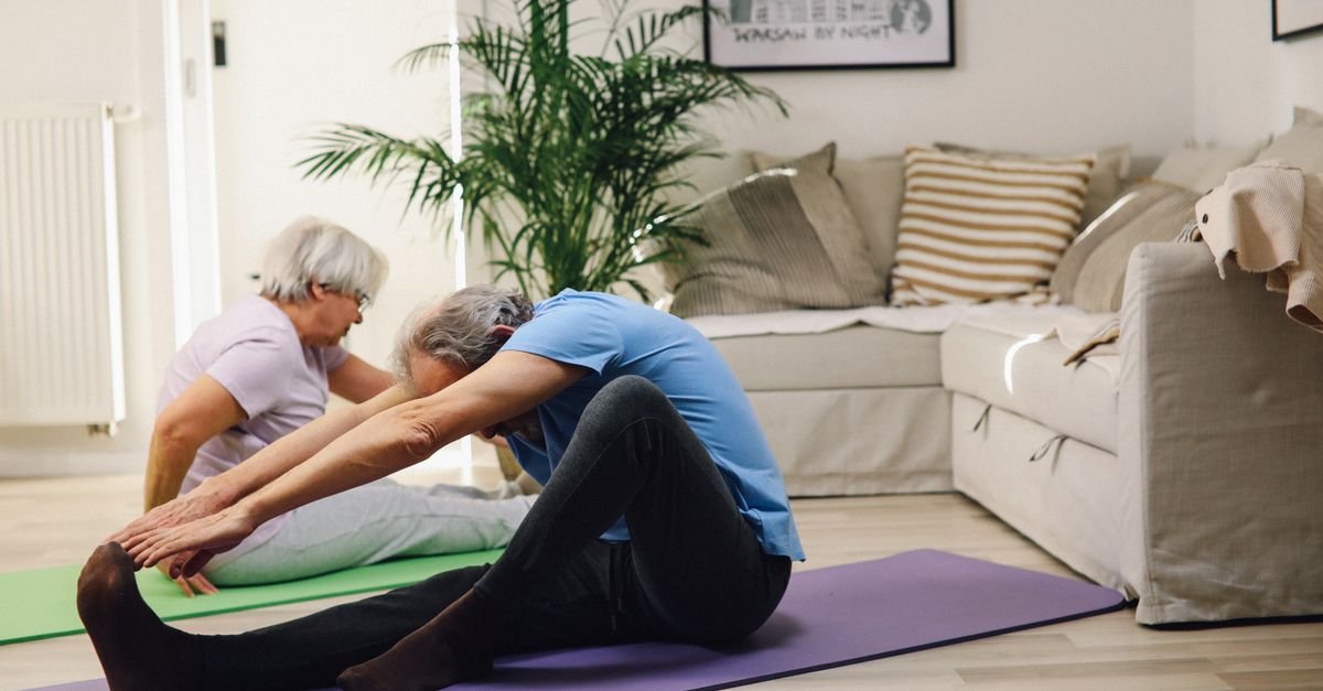 Two older adults stretch on exercise mats in a bright living room.