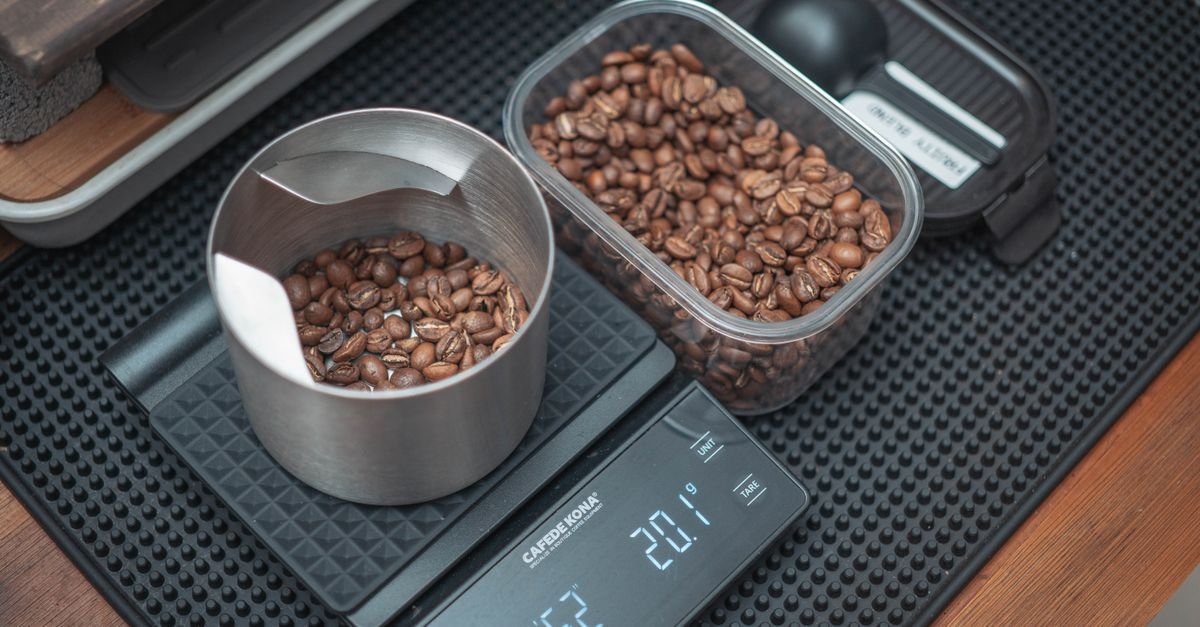 A metal measuring cup of coffee beans sits on a digital scale beside a clear container filled with more coffee beans.