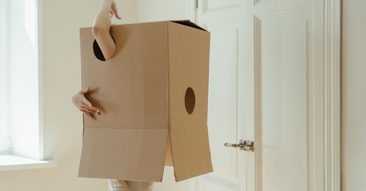 A person stands indoors wearing a large cardboard box with arm and leg openings.