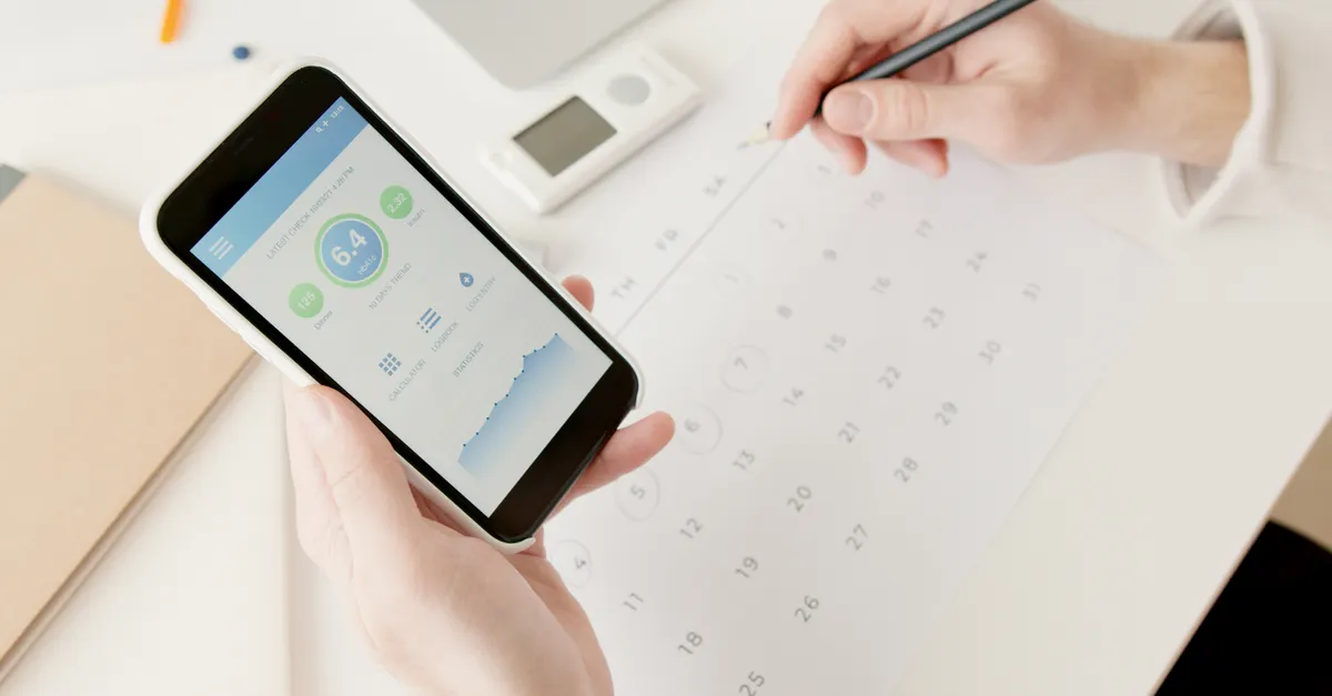 A person holds a smartphone displaying health metrics over a calendar, with a digital scale and pencil nearby.